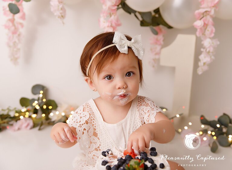 Little girl looking at camera with face covered in frosting while eating her first birthday cake - first birthday photographer jacksonville nc