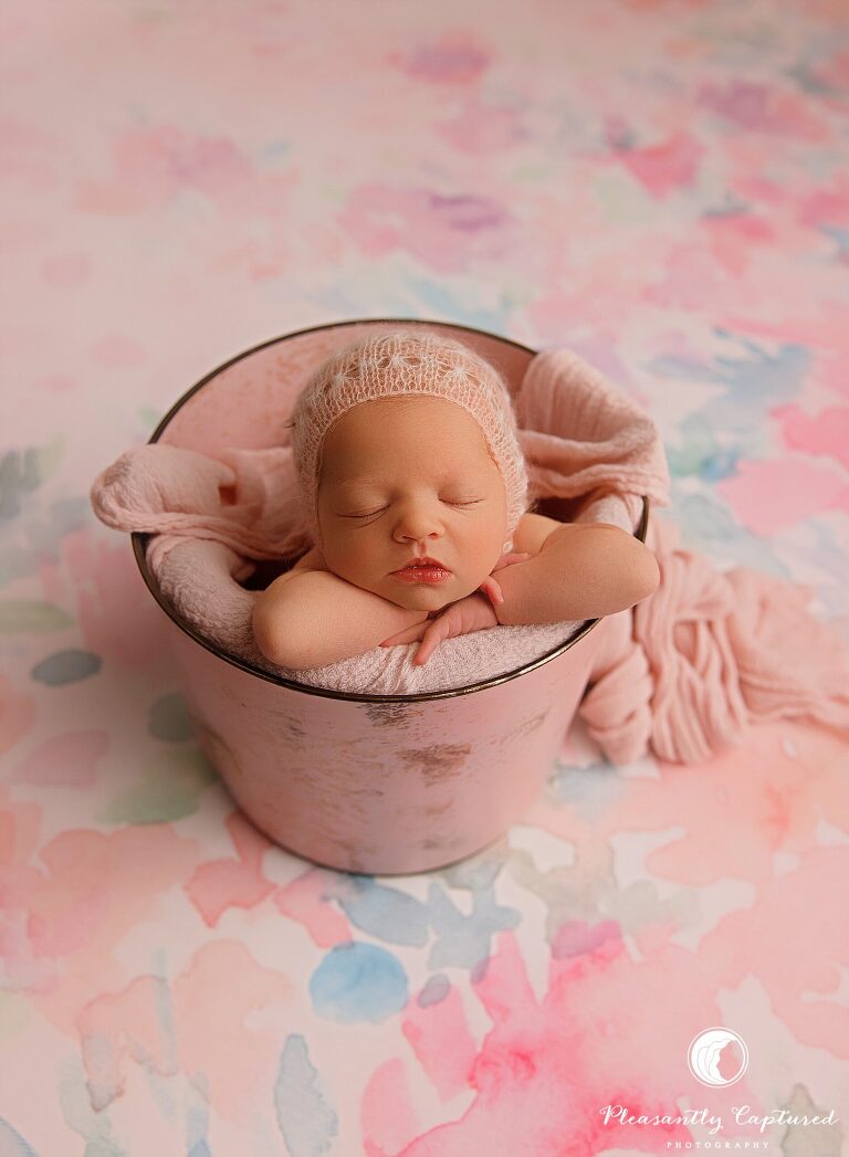 Baby girl in pink bonnet and wrap, inside bucket, on watercolor backdrop - Sneads Ferry Newborn Photography | New Family of 3