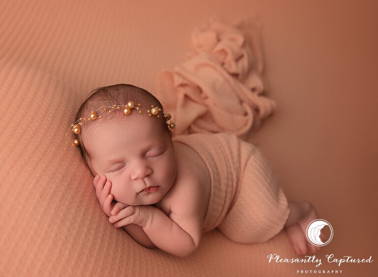 Newborn girl peacefully posed on a peach blanket with gold pearl headband during a studio session in Jacksonville, NC