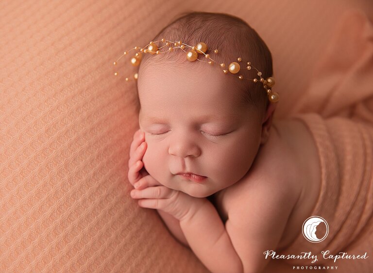 Sleeping newborn girl on peach textured blanket wearing a gold pearl headband in a timeless studio newborn portrait in Jacksonville, NC