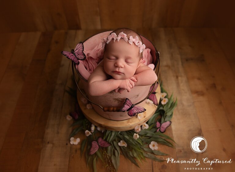 Newborn girl posed in a pink bucket surrounded by faux flowers and butterflies during whimsical studio newborn session in Jacksonville, NC