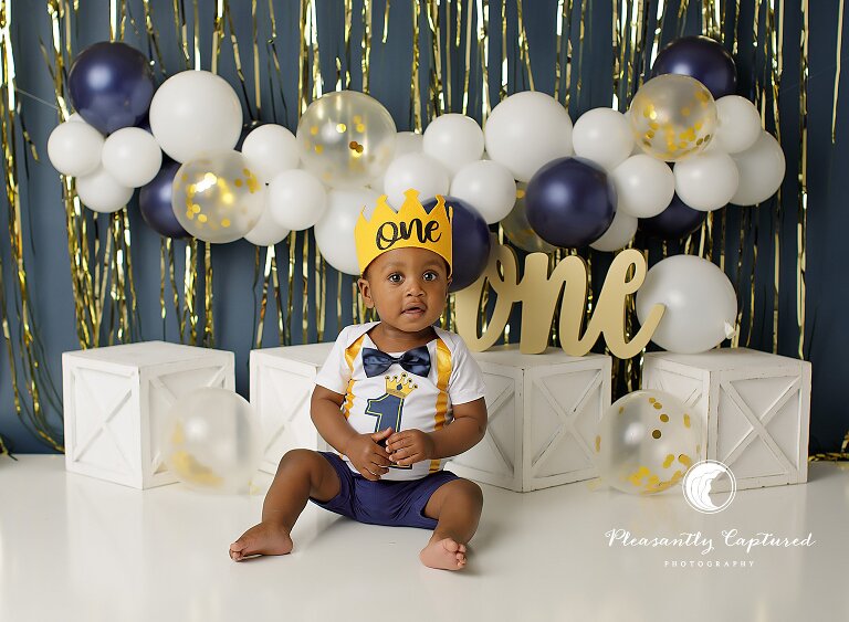 Baby boy sitting in a studio setup for a first birthday cake smash session with balloon garland and crown.