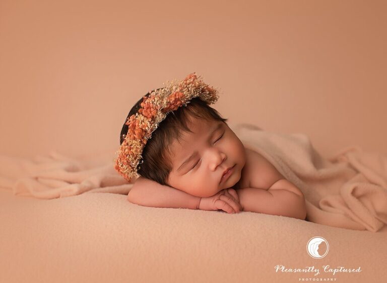 Baby girl sleeping on a peach backdrop with floral headpiece during a newborn mini session in Jacksonville, NC.